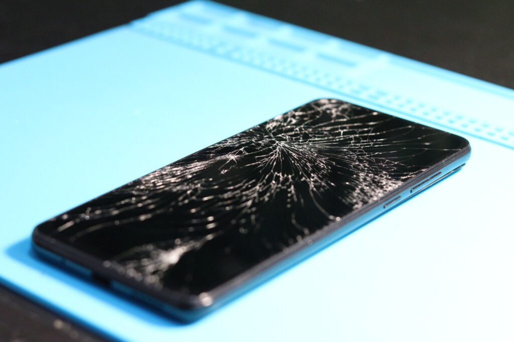 現在の画像: A cracked smartphone screen on a repair mat highlights technology mishaps.