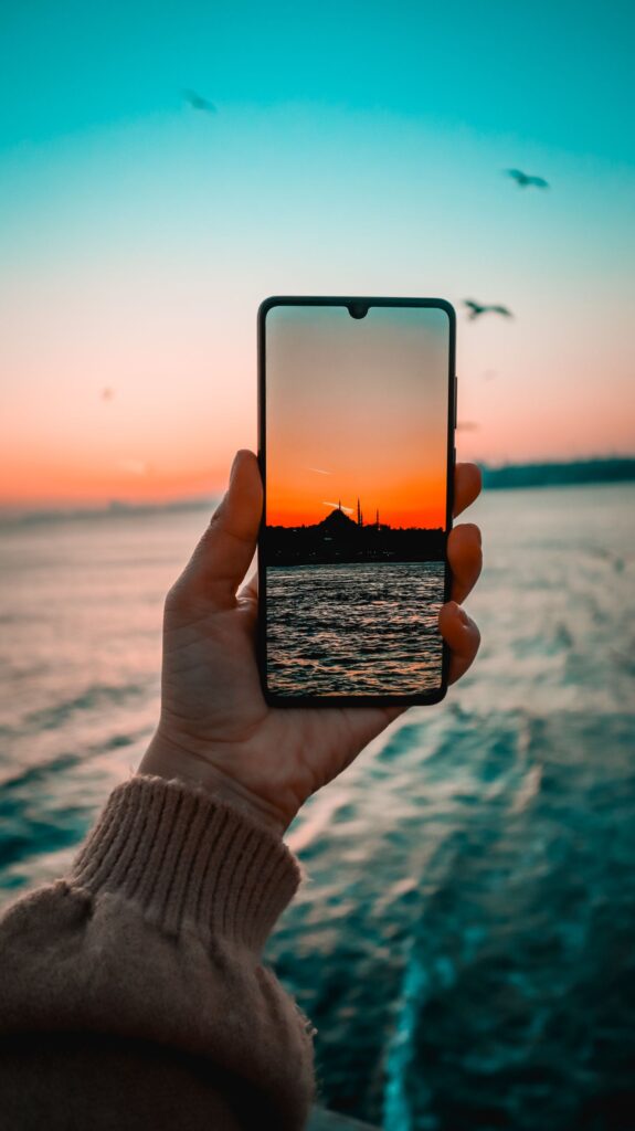 現在の画像: A hand holds a smartphone displaying a vibrant ocean sunset with birds flying overhead.