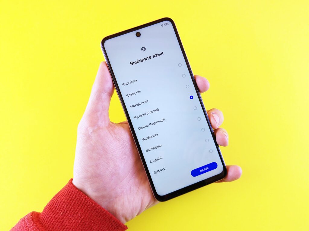 現在の画像: A hand in a red sleeve holding a smartphone displaying a language selection screen against a yellow background.