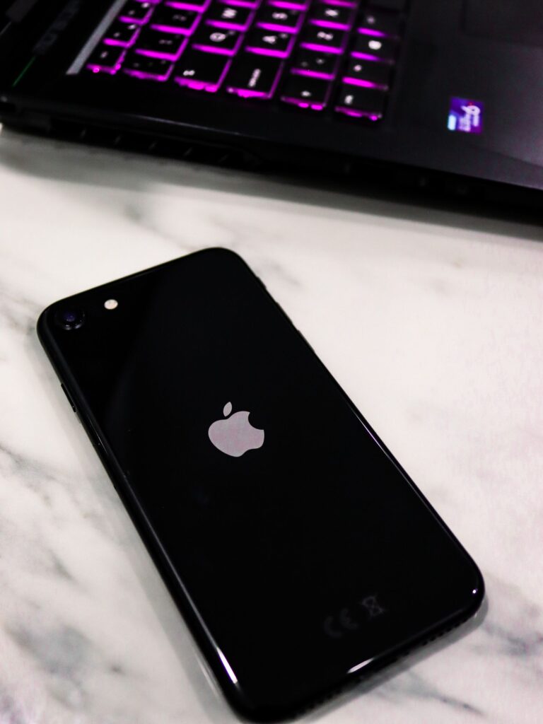 現在の画像: Close-up of a black smartphone with an apple logo on a marble surface next to a laptop with illuminated keys.