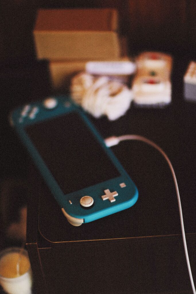 現在の画像: Close-up of a blue Nintendo Switch Lite console charging on a table with gaming accessories in a cozy indoor setting.