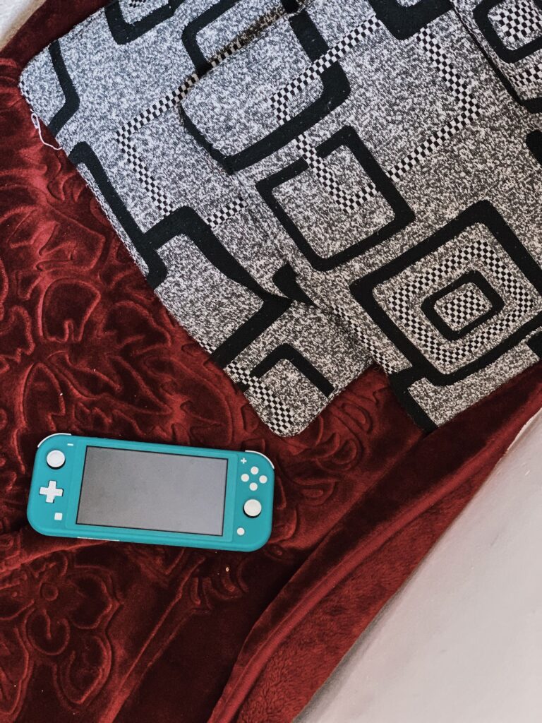 現在の画像: Close-up of a blue Nintendo Switch Lite console charging on a table with gaming accessories in a cozy indoor setting.