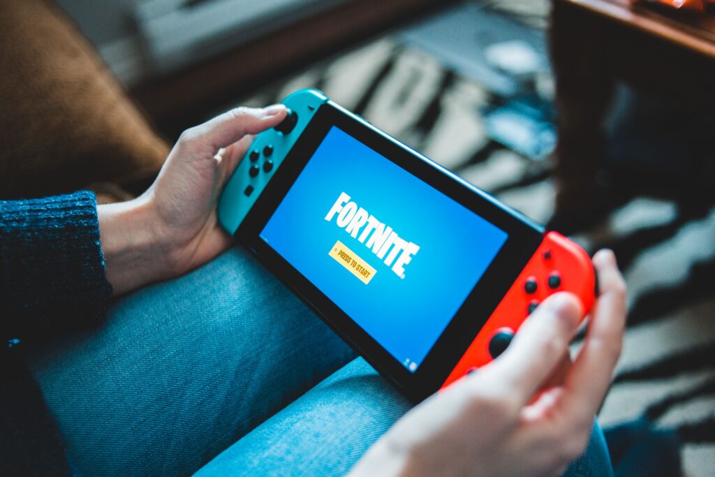 現在の画像: Close-up of hands holding a Nintendo Switch with Fortnite game screen indoors.