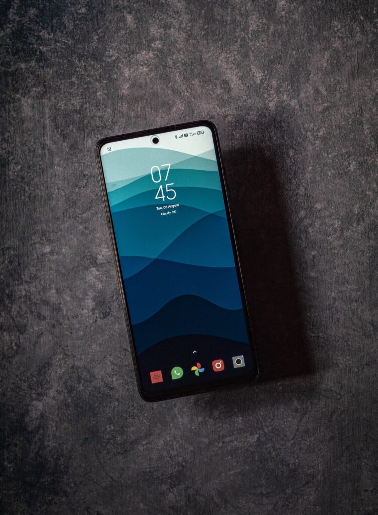 現在の画像: Elegant flatlay featuring a smartphone displaying 07:45 AM on a textured dark surface, emphasizing modern design.