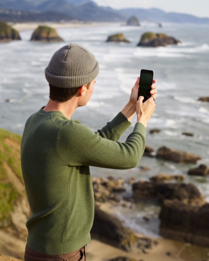 現在の画像: Side view of unrecognizable male traveler in hat and jumper looking at seascape and taking photo of vast sea surface on mobile phone